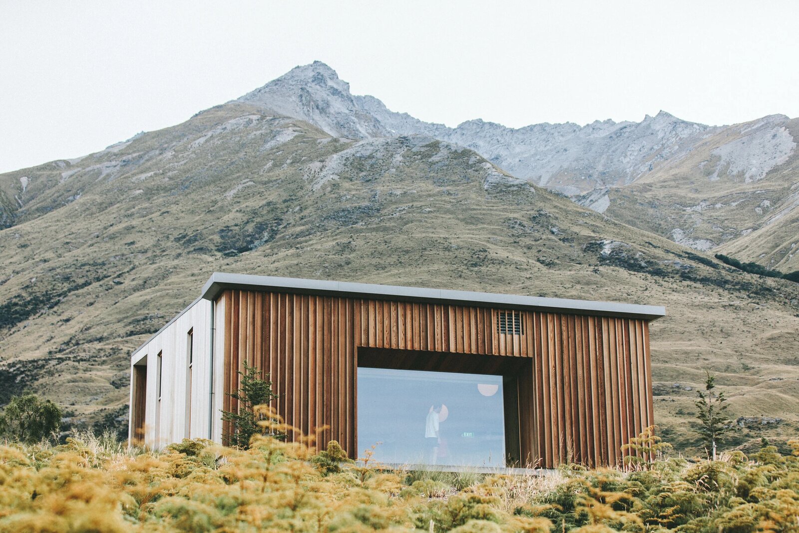 A modern wood-clad building set against a steep mountain under an overcast sky.